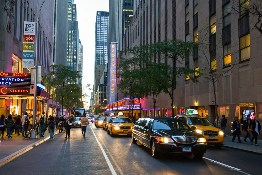 Busy city street at dusk with cars and pedestrians among tall buildings.